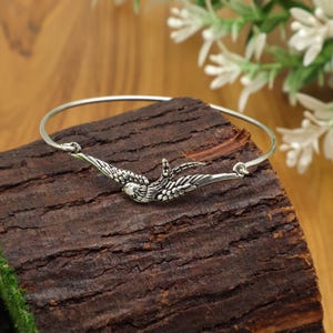 May include: A silver bangle bracelet with a bird motif. The bird is shown with its wings spread, and the bracelet has a simple, elegant design. The bracelet is displayed on a piece of dark wood.