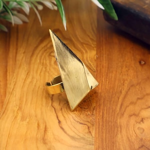 May include: A gold-colored statement ring with a geometric triangular design. The ring has a wide band and a large, textured triangle that extends upwards. The ring is set against a wooden surface.