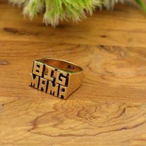 Op de afbeelding: Een goudkleurige ring met de woorden "BIG MAMA" in blokletters. De ring is rechthoekig met een platte bovenkant en zijkanten. De ring ligt op een houten oppervlak.