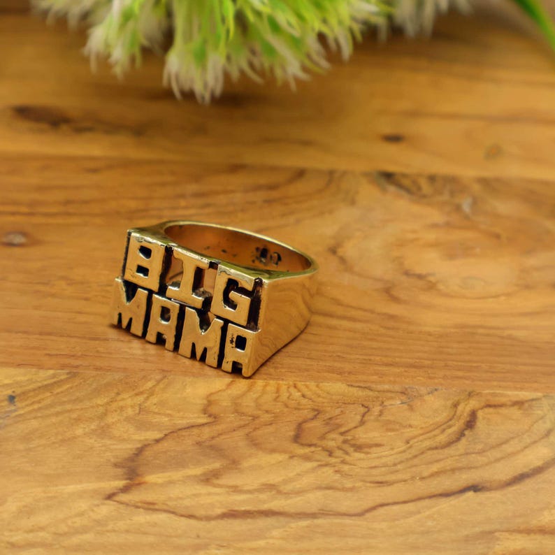 Op de afbeelding: Een goudkleurige ring met de woorden "BIG MAMA" in blokletters. De ring is rechthoekig met een platte bovenkant en zijkanten. De ring ligt op een houten oppervlak.