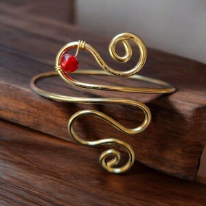 May include: A gold wire bracelet with a red bead accent. The bracelet is made of a single piece of wire that is twisted and shaped into a unique design.