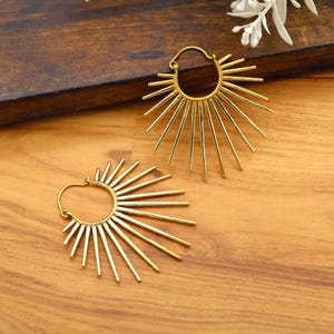 Op de afbeelding: Een paar goudkleurige oorringen. Elke oorring heeft een sunburst-ontwerp met uitstralende lijnen. De oorbellen worden gepresenteerd op een houten oppervlak, dat hun ingewikkelde ontwerp en metallic glans laat zien. Deze oorbellen zijn een stijlvol accessoire.