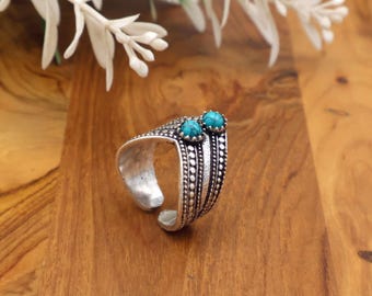 Peut inclure: Bague réglable argentée ornée de deux pierres turquoise rondes. La bague présente un motif détaillé avec des motifs pointillés et linéaires. La bague est présentée sur une surface en bois, avec des accents floraux blancs en arrière-plan.
