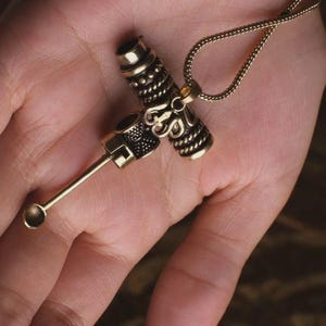 May include: A gold-colored pendant necklace featuring a detailed, ornate design. The pendant has a cross-like shape with intricate patterns and a small spoon-like attachment. The necklace chain is also gold and has a delicate, woven appearance.