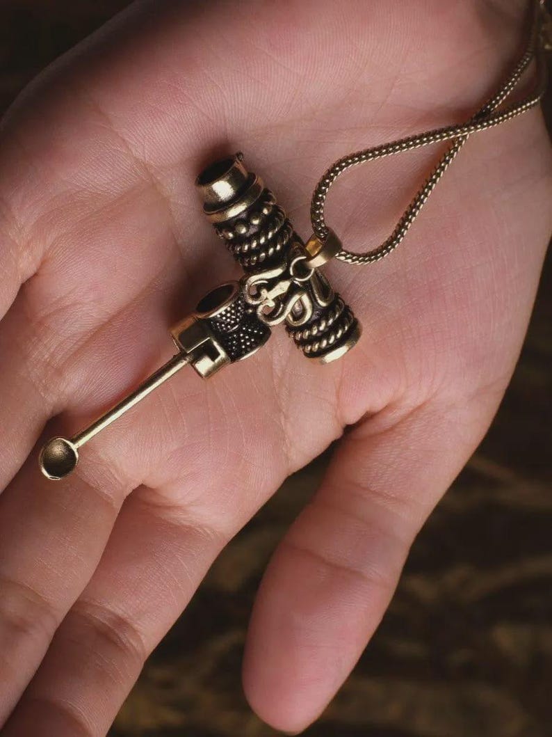 May include: A gold-colored pendant necklace featuring a detailed, ornate design. The pendant has a cross-like shape with intricate patterns and a small spoon-like attachment. The necklace chain is also gold and has a delicate, woven appearance.