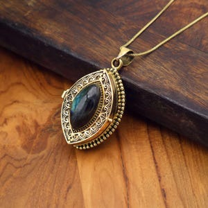 Può includere: Una collana con pendente dorato con un medaglione ovale. Il medaglione presenta un design a filigrana ornato e una pietra scura di forma ovale. La catena e il pendente dorati poggiano su una superficie di legno.