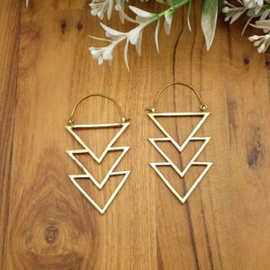 May include: A pair of gold-coloured earrings with a geometric design. Each earring features three stacked, open triangles suspended from a curved hoop. The earrings are displayed on a wooden surface, with white flowers in the background.