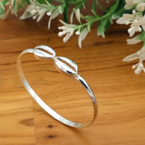 May include: A silver bangle bracelet with two cowrie shell charms. The bracelet has a simple, elegant design. The shells are detailed, adding a touch of coastal charm. The bracelet is suitable for everyday wear.