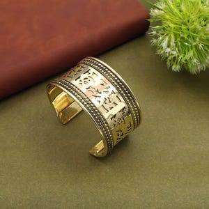 Peut inclure: Un bracelet manchette doré avec des motifs ajourés complexes. Le bracelet ouvert présente une large bande avec des motifs détaillés et une bordure texturée. Le bijou est présenté sur une surface verte avec un fond marron.