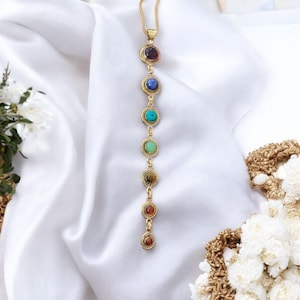 May include: A gold chain necklace with seven round gemstones in different colours. The stones are arranged in a vertical line, starting with a purple stone at the top and ending with a red stone at the bottom. The necklace is on a white background.