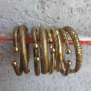 Op de afbeelding: Een set van acht gouden metalen armbanden met verschillende texturen en patronen. De armbanden zijn op elkaar gestapeld.