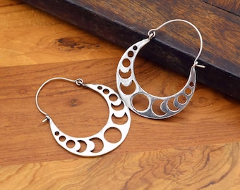 May include: A pair of silver-colored hoop earrings. Each earring features a crescent moon shape with a series of circular cutouts. The earrings are displayed on a wooden surface, showcasing their intricate design and metallic sheen.