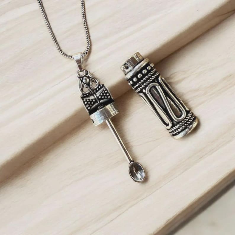 May include: A silver-colored pendant necklace with a decorative cylindrical container and a small spoon. The container is open, revealing the spoon. The necklace and container are on a light-colored wooden surface.