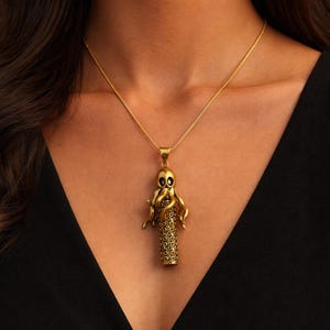 May include: Gold-coloured octopus pendant necklace. The pendant has an octopus design with detailed tentacles and a cylindrical body. The necklace is a delicate chain, and the pendant is suspended from it. The necklace is worn against a black background.
