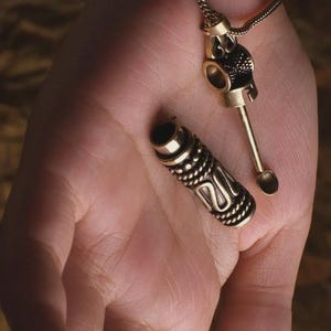 May include: A gold-colored metal pendant necklace with a small spoon and a cylindrical container. The container is detached and has a detailed, textured design. The necklace is displayed on a hand.