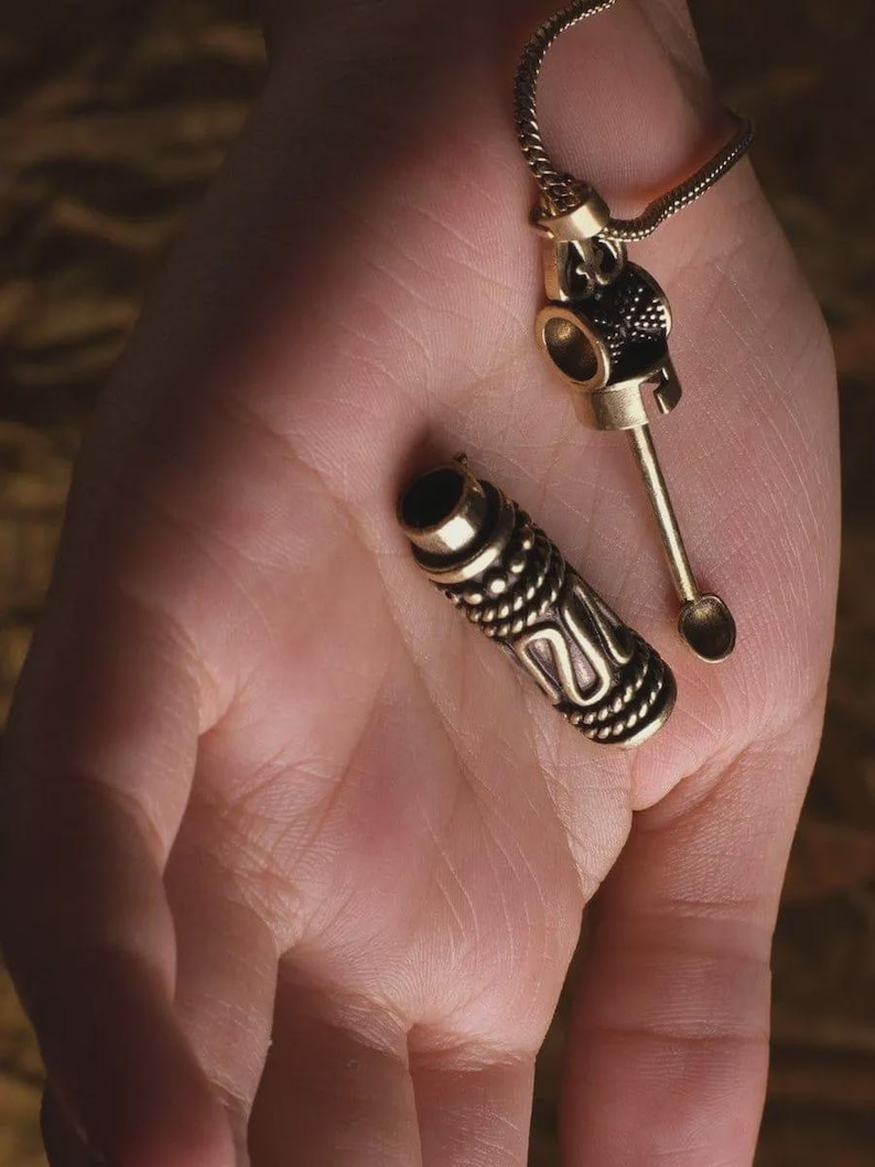 May include: A gold-colored metal pendant necklace with a small spoon and a cylindrical container. The container is detached and has a detailed, textured design. The necklace is displayed on a hand.