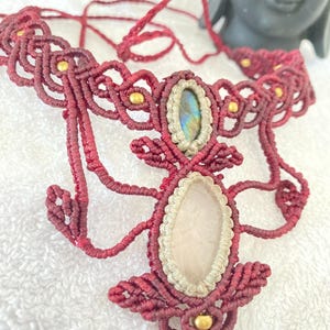 May include: A burgundy macrame necklace with an oval, iridescent gemstone and a larger, oval, light pink gemstone. The necklace has gold beads and intricate knotwork, creating a bohemian style. The necklace is made with a macrame technique.