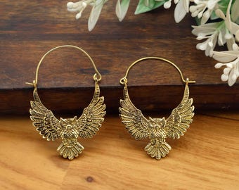 May include: A pair of gold-colored hoop earrings. Each earring features a detailed owl design with outstretched wings. The earrings are set against a wooden surface with white flowers in the background.