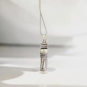May include: Silver-toned pendant necklace with an ornate, cylindrical design. The pendant features intricate patterns and a decorative top, suspended from a delicate chain. The necklace is displayed against a white background, highlighting its detailed craftsmanship.