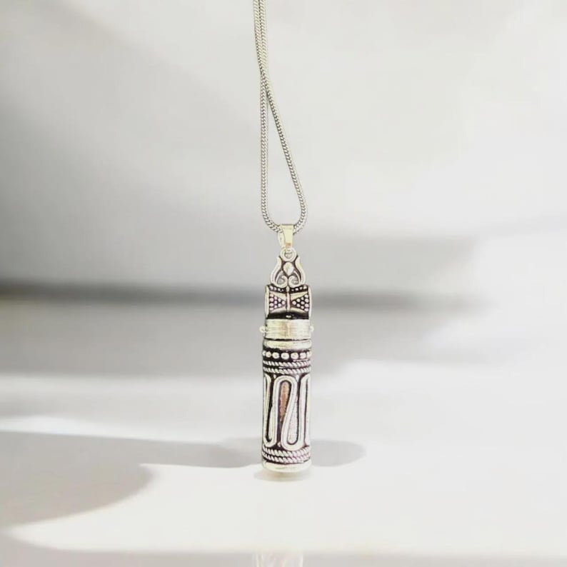 May include: Silver-toned pendant necklace with an ornate, cylindrical design. The pendant features intricate patterns and a decorative top, suspended from a delicate chain. The necklace is displayed against a white background, highlighting its detailed craftsmanship.