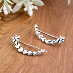 May include: A pair of silver ear cuffs with a flower design and a row of hearts. The cuffs are curved and have a delicate, feminine look.