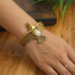 Könnte beinhalten: Ein goldfarbenes Metallarmband in Form eines Vogels mit ausgebreiteten Flügeln. In der Mitte des Vogels befindet sich ein ovaler, schillernder weißer Stein. Die Metalloberfläche weist detaillierte, geätzte Linienstrukturen auf den Flügeln auf.