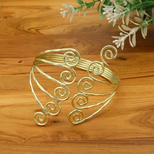 May include: Gold-coloured arm cuff with a hammered texture and decorative swirl design. The cuff features multiple bands and stylized spiral accents, creating an elegant accessory. The cuff is approximately 15 cm in diameter.