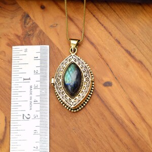 May include: A gold-colored pendant necklace with an oval-shaped, iridescent gemstone. The pendant is approximately 2 inches long and features intricate filigree detailing. The necklace is displayed on a wooden surface.