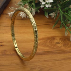 Puede incluir: Un brazalete circular de tono dorado que presenta un patrón en relieve continuo y repetitivo de pequeñas escamas semicirculares superpuestas en toda su superficie. La banda de metal tiene un acabado pulido que refleja la luz en su borde.