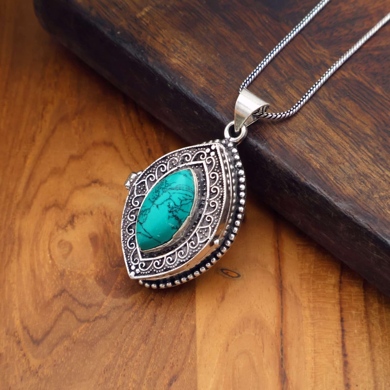 May include: A silver pendant necklace with a turquoise stone center. The pendant has an ornate, detailed design around the stone. The necklace chain is a thin, silver chain. The pendant is resting on a wooden surface.