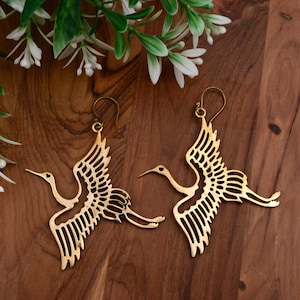 May include: A pair of gold-colored crane earrings. The earrings are intricately designed with openwork wings and a graceful, elongated neck. The earrings are displayed on a wooden surface with greenery.