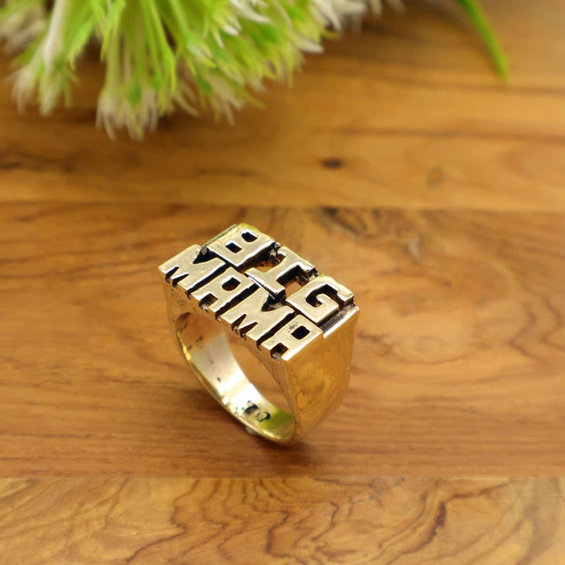 Op de afbeelding: Een goudkleurige ring met de woorden "BIG MAMA" in blokletters. De ring heeft een rechthoekig ontwerp en ligt op een houten oppervlak. Een opvallend sieraad.
