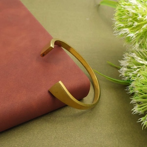 Op de afbeelding: Een goudkleurige manchetarmband met een vierkante opening. De armband ligt op een bordeauxrood oppervlak met een groen-witte bloemstuk op de achtergrond. De armband heeft een gladde, gepolijste afwerking.