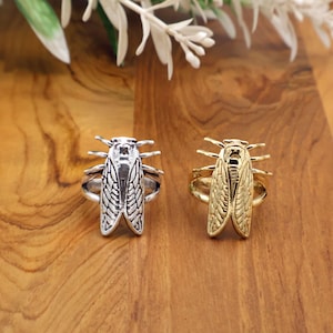 May include: Two insect-themed rings. One ring is silver-colored, the other is gold-colored. Both rings feature a detailed cicada design with wings and body. The rings are displayed on a wooden surface.