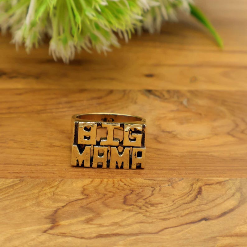 Op de afbeelding: Een goudkleurige ring met de woorden "BIG MAMA" in blokletters. De ring staat tegen een houten achtergrond, met een wazig groen en wit bloemelement op de achtergrond.