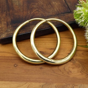 Può includere: Due orecchini a cerchio color oro. Gli orecchini sono circolari e hanno una superficie liscia e lucida. Sono appoggiati su una superficie di legno. Gli orecchini sono di un colore oro brillante.