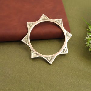 May include: A silver-colored bangle bracelet with a starburst design. The bracelet features triangular points around the outer edge, each with etched linear patterns. The bracelet is circular and rests on a brown and green surface.