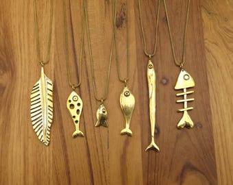 May include: A collection of gold-colored pendant necklaces featuring various designs, including a feather, fish, and fish skeleton. The pendants hang on delicate chains, displayed against a wooden background.