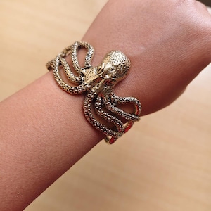 Pode incluir: Pulseira dourada de polvo. A pulseira envolve o pulso, com a cabeça e os tentáculos do polvo formando o design. A pulseira é feita de metal brilhante e tem uma textura detalhada. É uma joia única.