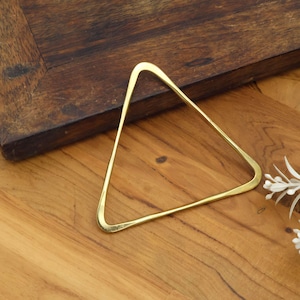 May include: A gold-colored, triangular bangle bracelet with rounded corners. The bracelet is made of a shiny metal and is displayed on a wooden surface. The bangle's design is simple and modern, suitable for everyday wear.