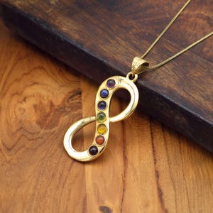 Op de afbeelding: Goudkleurige ketting met een oneindigheidssymbool. De hanger heeft zeven kleurrijke edelstenen, die de chakra's vertegenwoordigen. De ketting ligt op een houten oppervlak.