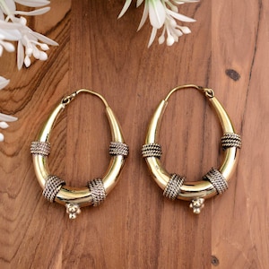 May include: A pair of gold-colored hoop earrings with a teardrop shape. The earrings have decorative bands of twisted metal and small floral accents. The earrings are displayed on a wooden surface with white flowers in the background.
