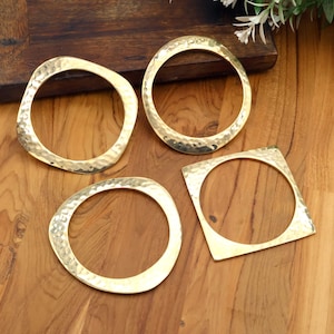 May include: Four gold-colored bangles with a hammered texture. The bangles are in various shapes: a circle, a square, and two irregular shapes. The bangles are displayed on a wooden surface.