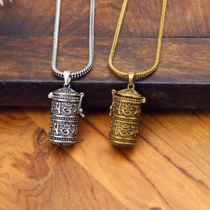 Puede incluir: Dos collares con colgantes cilíndricos. Uno es plateado, el otro dorado. Ambos colgantes tienen texto grabado y están suspendidos de collares de cadena a juego. Los colgantes están sobre una superficie de madera.