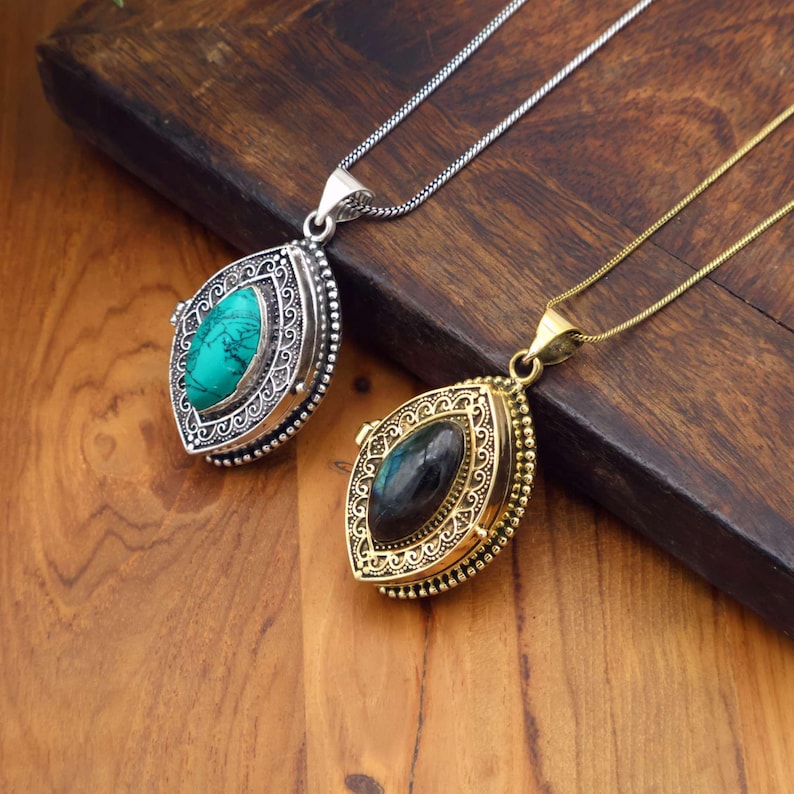 May include: Two ornate pendant necklaces. One features a silver-toned setting with a turquoise stone, the other a gold-toned setting with a dark stone. Both have intricate designs and hang on delicate chains, displayed on a wooden surface.