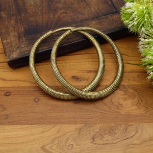 May include: Two gold-colored, textured bangles are displayed on a wooden surface. The bangles are circular, with a detailed, dotted pattern. The background includes a dark wooden box and a sprig of white flowers.