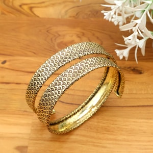 May include: Two gold-coloured bangles with a detailed, repeating pattern. The bangles are made of metal and have a slightly aged appearance. The design features small, curved shapes, creating a textured surface. The jewellery is displayed on a wooden surface.