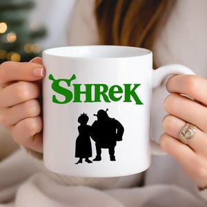 May include: White ceramic mug with a green "Shrek" logo and a black silhouette of Shrek and Fiona.