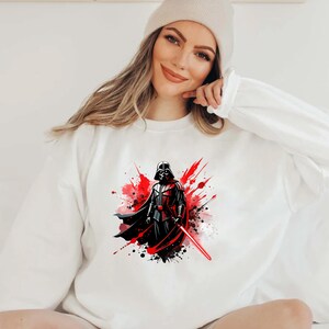 Op de afbeelding: Wit sweatshirt met een afbeelding van Darth Vader, een rood lichtzwaard en een rood en zwart verfspatterontwerp. De Star Wars-thema sweatshirt heeft een ronde hals en lange mouwen.