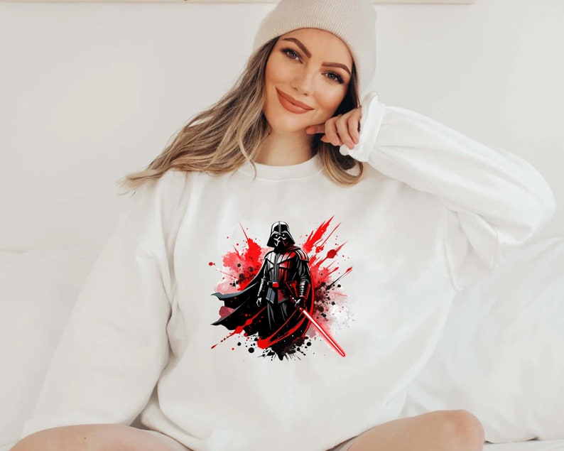 Op de afbeelding: Wit sweatshirt met een afbeelding van Darth Vader, een rood lichtzwaard en een rood en zwart verfspatterontwerp. De Star Wars-thema sweatshirt heeft een ronde hals en lange mouwen.
